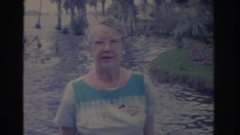 1962 Florida home-movie frame showing an adult woman facing the camera with cypress swamp water and shoreline greenery behind her.