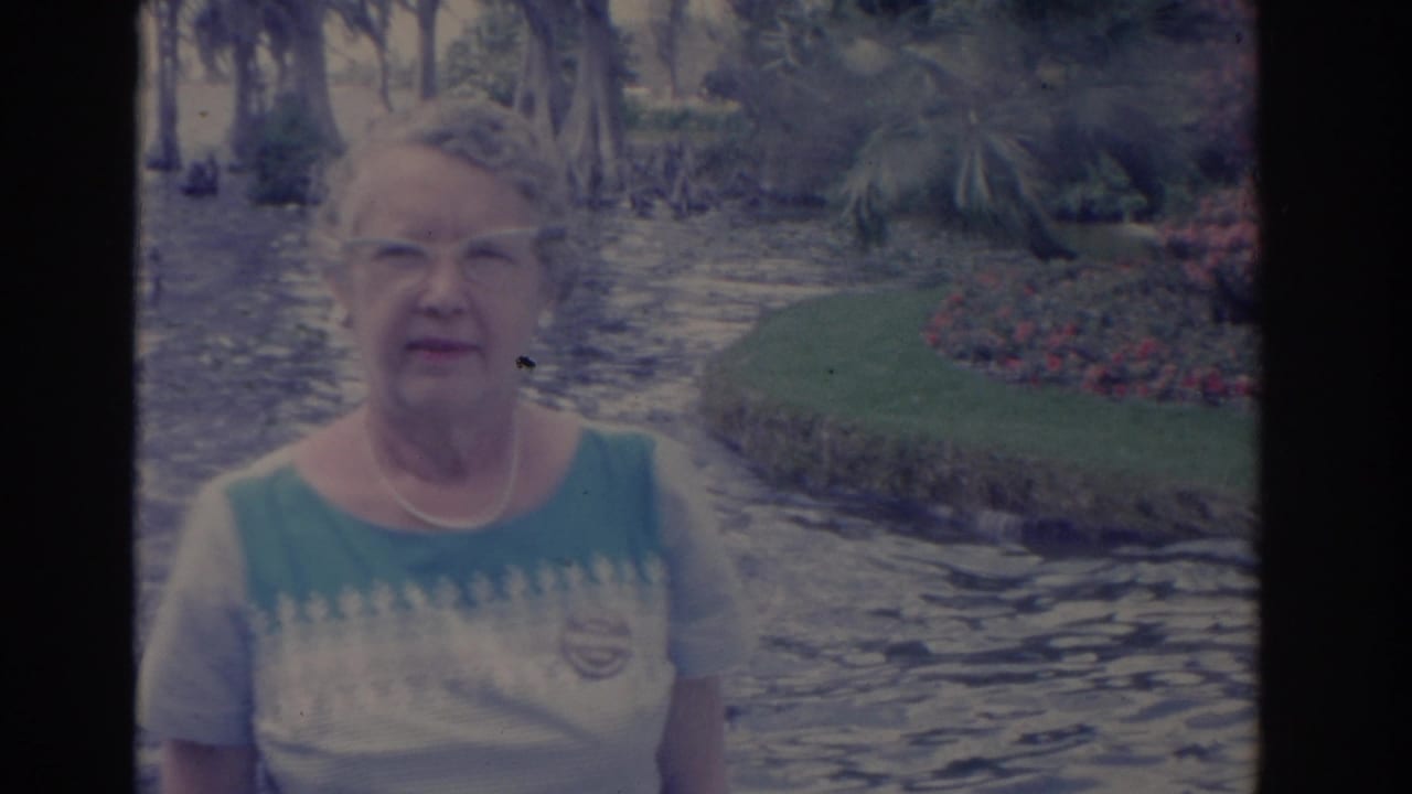 Elderly woman with glasses standing beside flowing water in a garden.