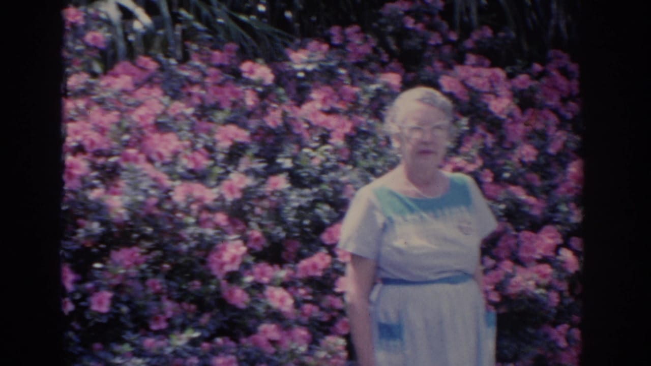 An elderly woman in a light dress and glasses poses in front of vivid pink flowers in the 1962 Florida garden footage.