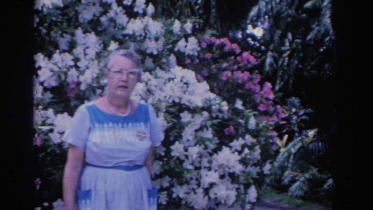 1962 Florida home-movie frame of an adult woman in a light dress standing before white and pink flowering shrubs and dark palm foliage.