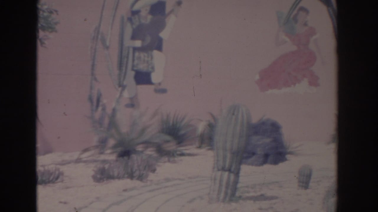 Late frame from the 1962 Florida 8mm pan showing cacti, rocks, sandy ground, and painted mural figures on a pink stucco wall.