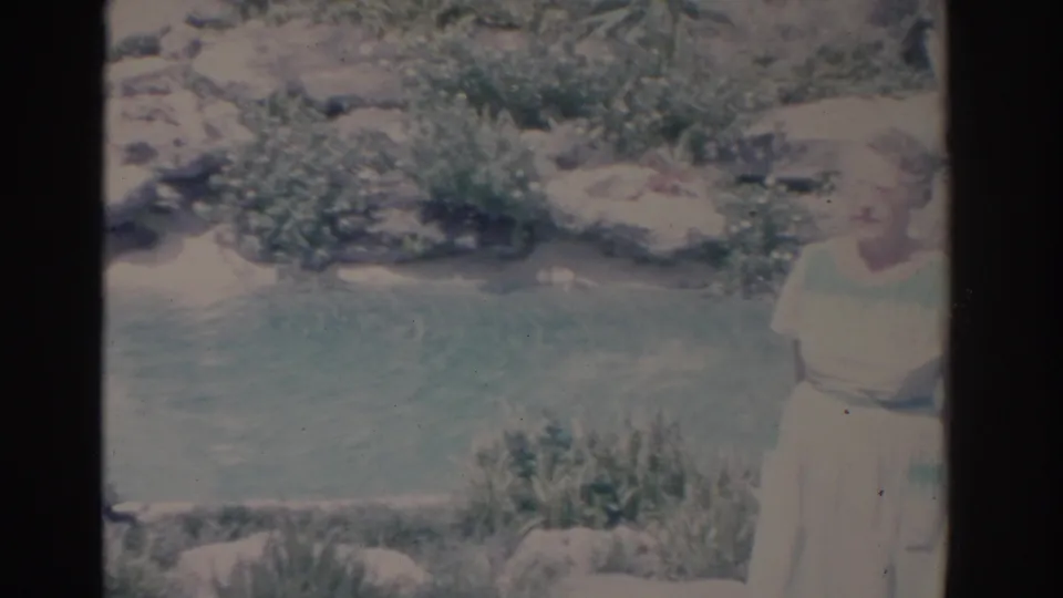 1962 Florida home-movie frame with a woman in a light dress standing beside a rocky river or garden pool.
