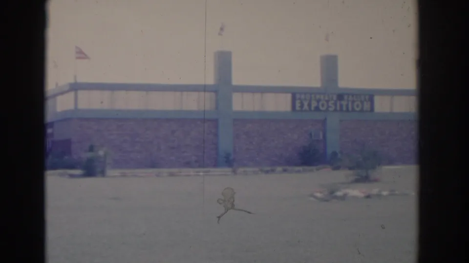 1962 Florida 8mm frame showing an exposition building exterior with blue columns, brick facade, flags, and landscaped pavement.