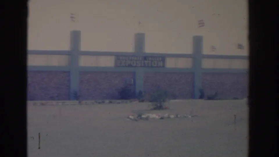 1962 Florida home-movie view of the exposition building sign and facade, seen across open pavement with small flags above.