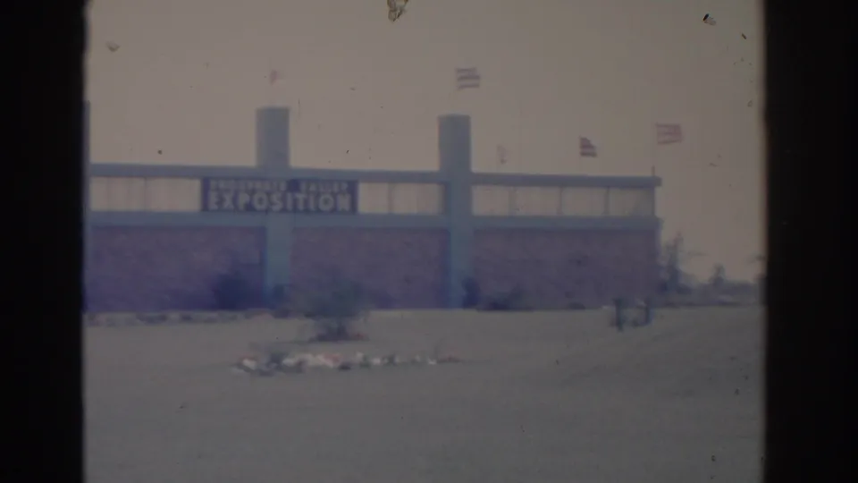 1962 Florida 8mm frame of the exposition building from farther left, with the sign, flags, pavement, and landscaped bed visible.