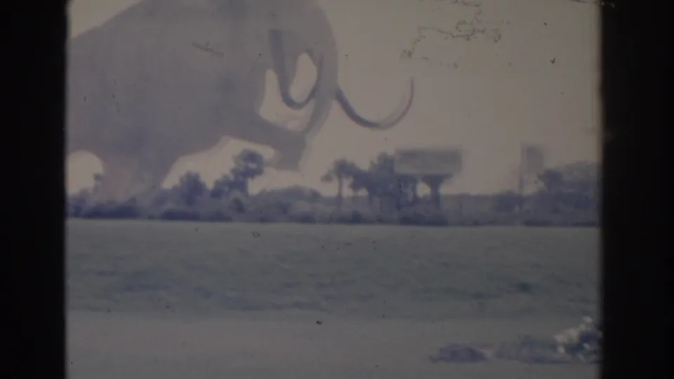 Faded 1962 Florida 8mm view of the mammoth statue and tusk over a flat lawn, with palms, shrubs, and roadside signage beyond.