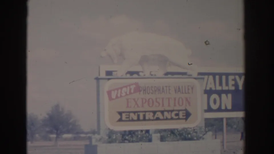 1962 Florida frame showing a Phosphate Valley Exposition entrance sign with an animal figure on top, nearby cars, shrubs, and faded color.