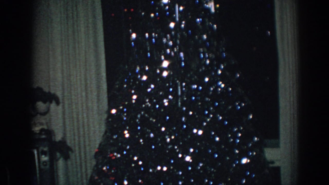 Lit Christmas tree in front of curtains and a window inside a dim room.