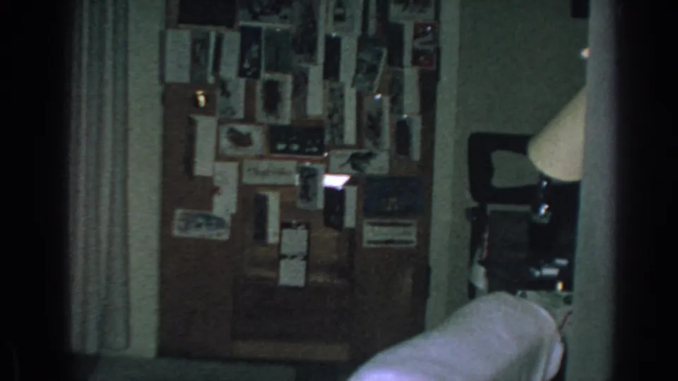 1962 Gary, Indiana home-movie frame showing a low-lit private room with a dense photo wall, curtain, couch edge, and lamp.