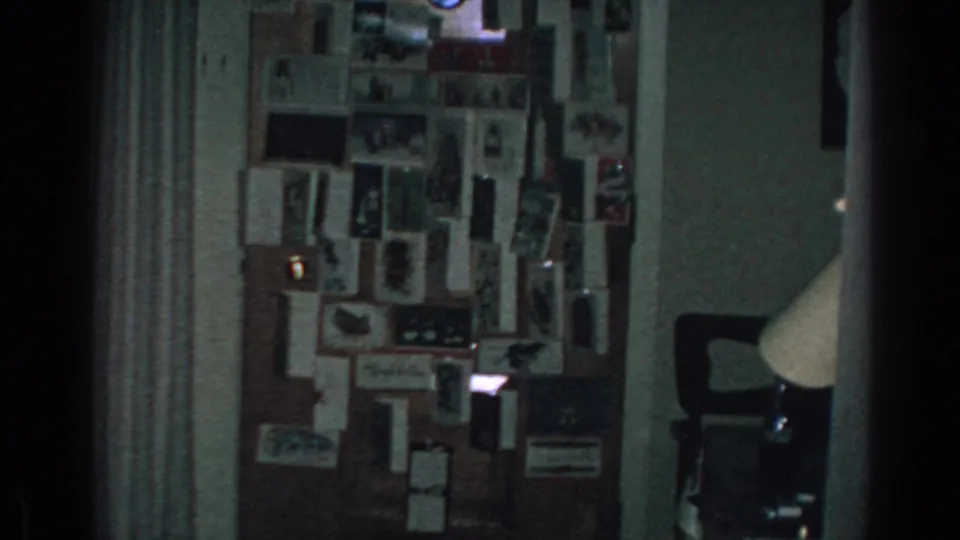 The 8mm frame holds on the same Gary, Indiana interior, with a dense wall collage and lamp partially visible to the right.