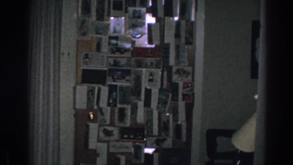 A low-lit 1962 home-movie frame shows a private interior wall densely covered with photographs, postcards, and clipped papers.