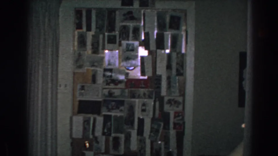 1962 Gary, Indiana home-movie frame showing a dim room wall with a dense display of cards and photographs.