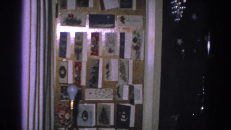 1962 Gary, Indiana home-movie frame showing the decorated wall board beside curtains and faint Christmas lights in a dark room.