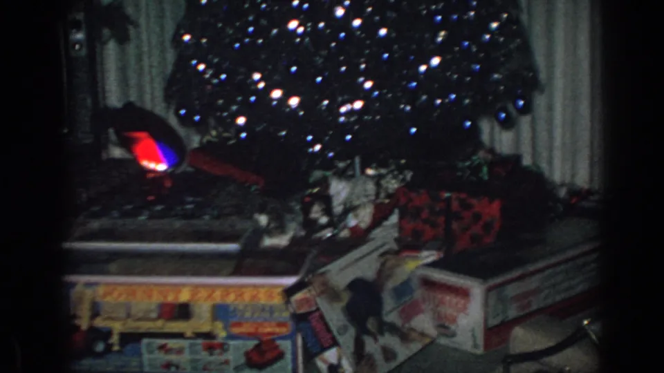 1962 Gary, Indiana home-movie frame showing reflective Christmas decorations, wrapped presents, toy boxes, and curtains in a private living room.