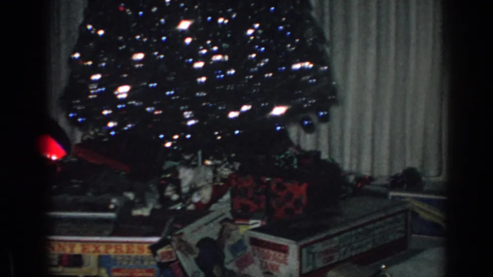 1962 Gary, Indiana home-movie frame with a decorated Christmas tree, glowing ornaments, curtains, and toy boxes grouped around the base of the tree.