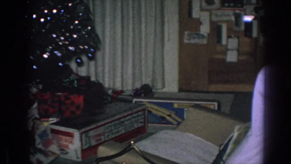1962 Gary, Indiana home-movie frame showing a dim living room corner with a Christmas tree, presents, open boxes, curtains, and a wall bulletin board.