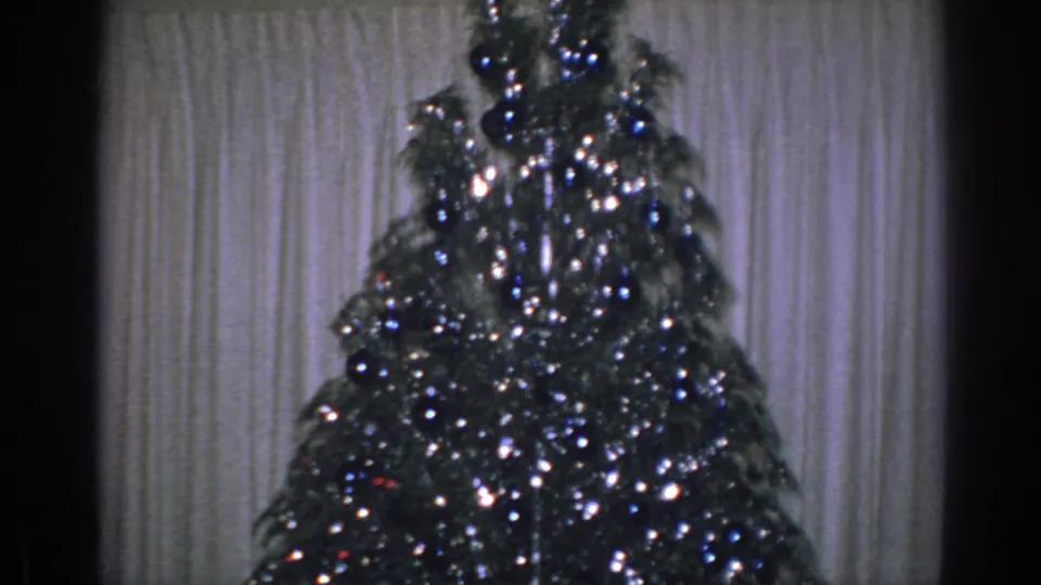 1962 Gary, Indiana home-movie frame with a tinsel-covered Christmas tree standing in front of pale curtains, framed by dark 8mm film edges.