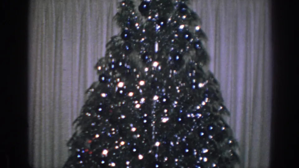 1962 Gary, Indiana home-movie frame showing a closer view of the tinsel-covered Christmas tree, ornaments, and pale curtains in a private interior.