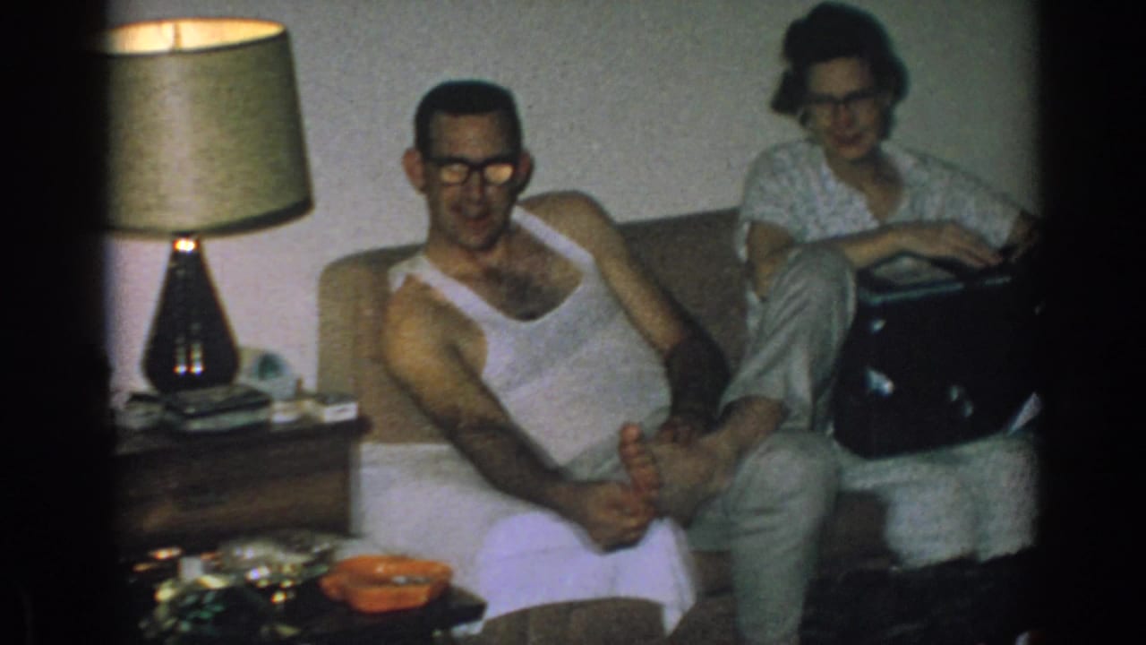 1962 Gary, Indiana home-movie frame of two adults with glasses seated on a couch, with a table lamp and small black box nearby.