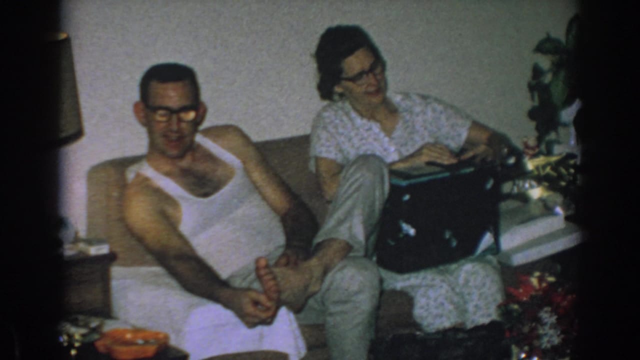 Couple sits on a couch while the woman rests a black box on her lap near floral decor.