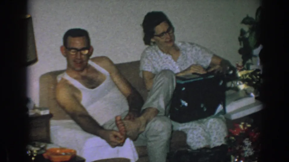 1962 Gary, Indiana frame showing a relaxed couple on a couch, the woman holding a black box with flowers and living-room furniture visible.