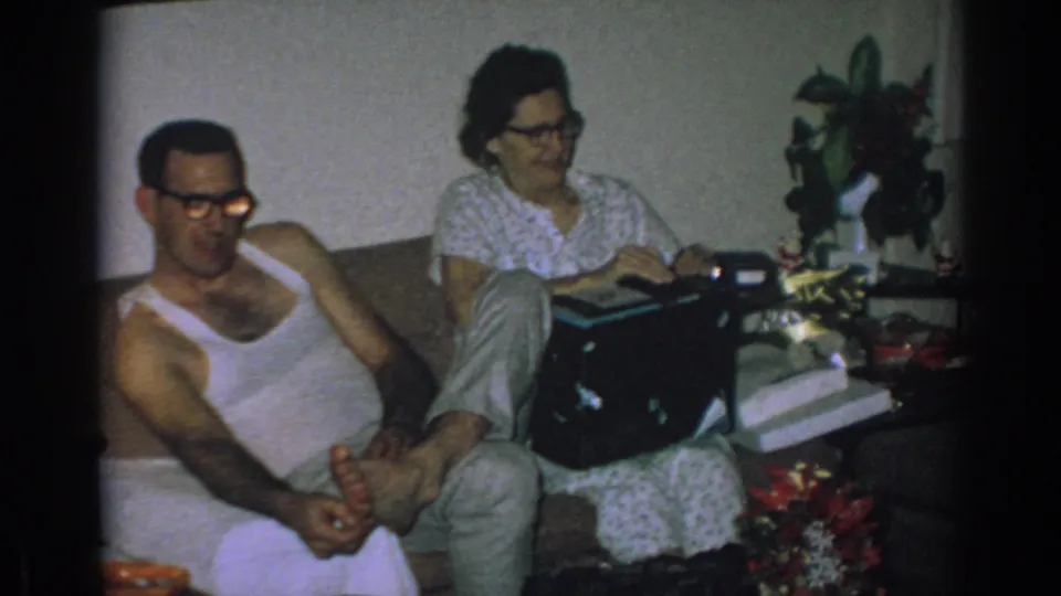 Home-movie frame from 1962 Gary, Indiana showing a relaxed adult couple on a couch, with the woman holding a compact black box.
