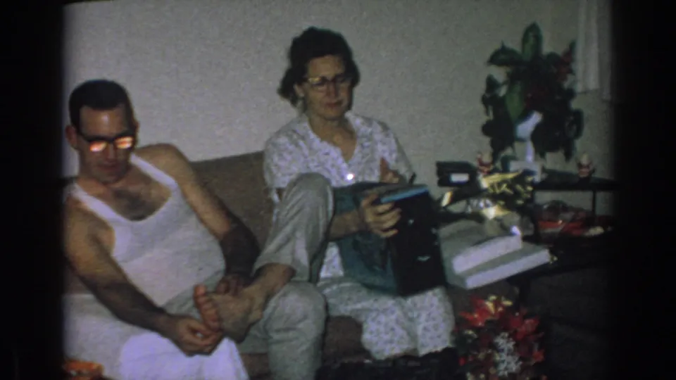 1962 Gary, Indiana interior frame with a woman examining a black box on her lap as a man sits beside her in a dim living room.