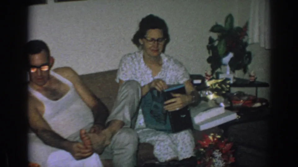 In a 1962 Gary, Indiana living room, a woman in glasses opens a gift on a couch while a man in a white undershirt sits beside her.