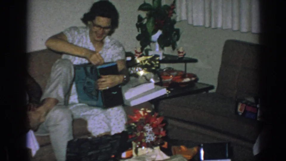 The woman leans over the gift on her lap, with a living-room chair, side table, plant, and wrapped boxes visible around her.