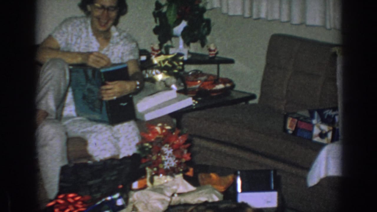 Woman with glasses opens a Christmas gift beside presents in a living room.