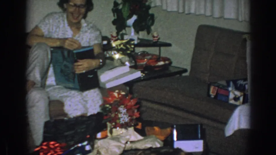 In a 1962 Gary, Indiana home-movie frame, a woman with glasses opens a gift near wrapped presents, a couch, and holiday decor.