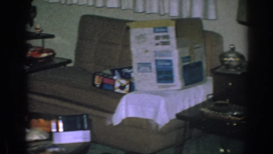 A 1962 Gary, Indiana living room frame shows a couch holding a large appliance box, with wrapped gifts, side tables, and curtains nearby.