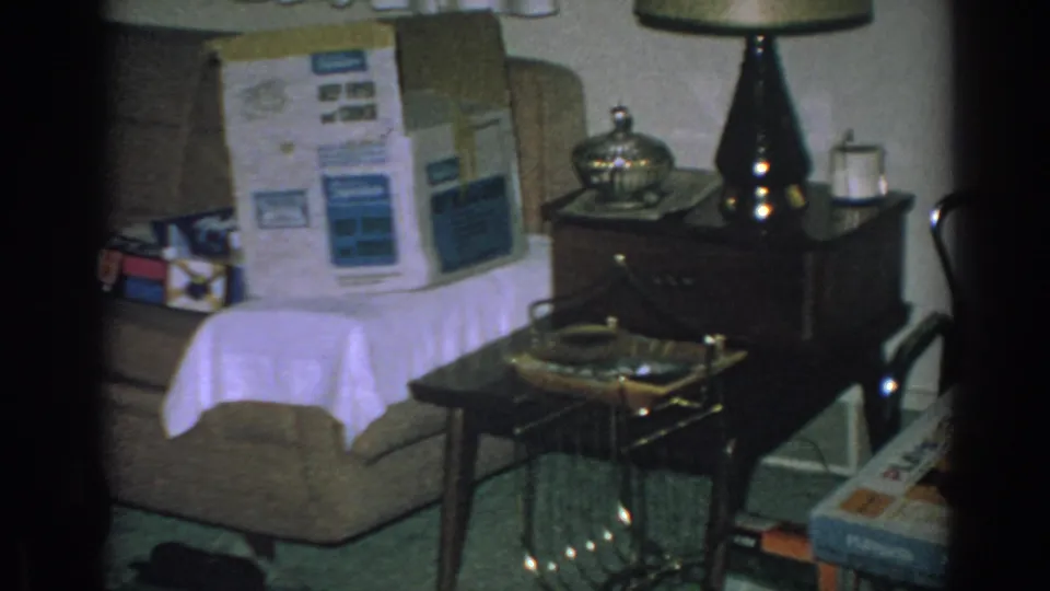 The frame shows a Gary, Indiana living room with a couch, large appliance box, dark table lamp, decorative dish, wrapped gifts, and small tables.