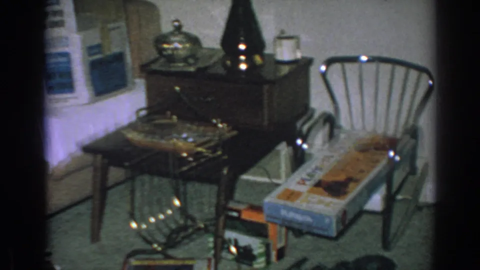 A 1962 home interior frame shows a side table with lamp, a metal-framed chair, appliance box, wrapped gifts, and small household tables.