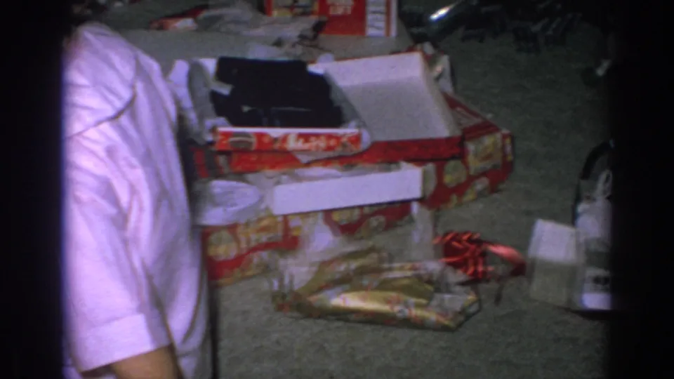 1962 home-movie frame in a private Gary, Indiana interior with opened boxes, wrapping, carpet, and a partially visible adult torso.