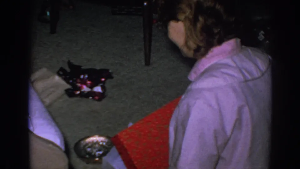 A woman sits on the carpet among red wrapping paper, an open package, and household items during the 1962 Christmas scene.