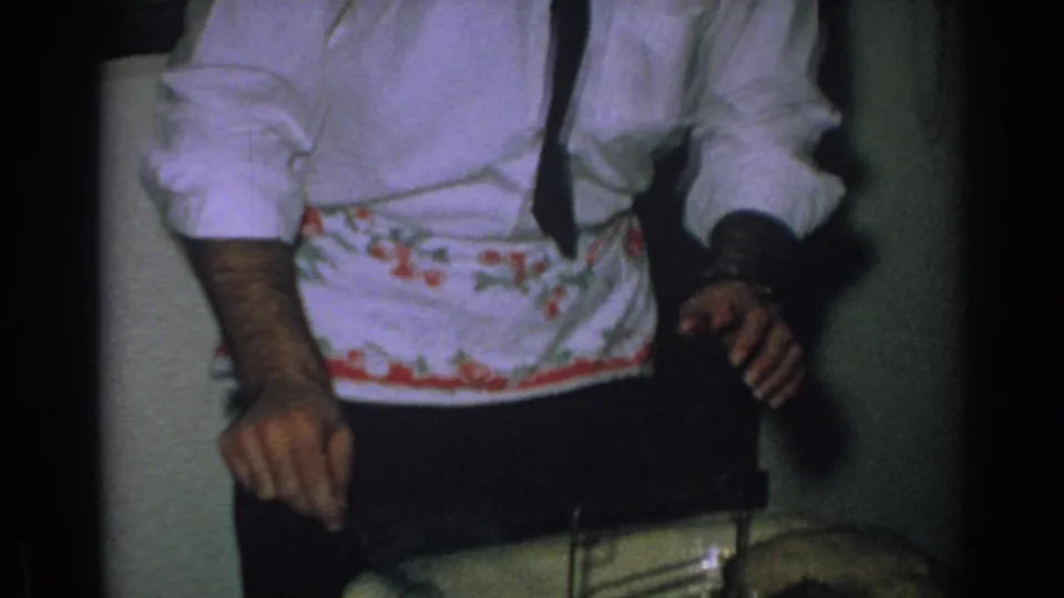 A man in a white shirt, dark tie, and floral apron stands in a private interior during the 1962 Gary, Indiana holiday gathering.