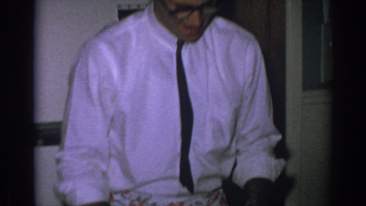 Man in white shirt and dark tie looks down while wearing a floral apron indoors.