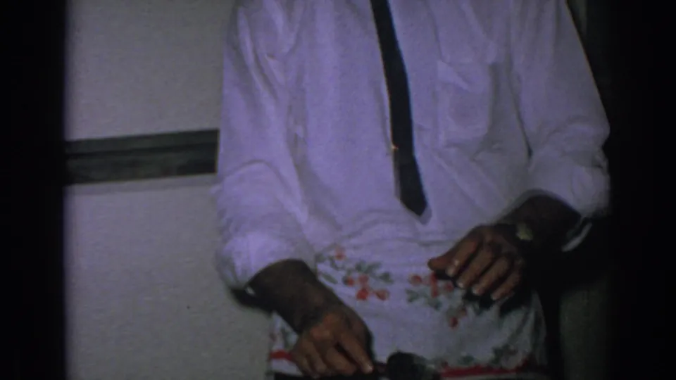 A man in a white shirt, dark tie, and floral apron works with a utensil near a dish in a dim Gary, Indiana kitchen scene.