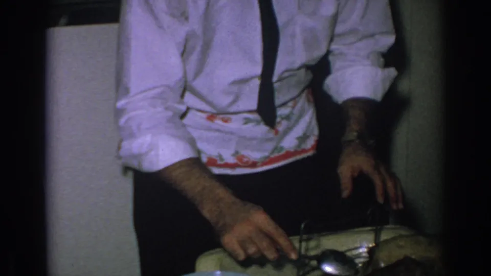 The man in formal shirt and tie reaches toward a casserole or serving dish with a spoon during a 1962 home-kitchen moment.