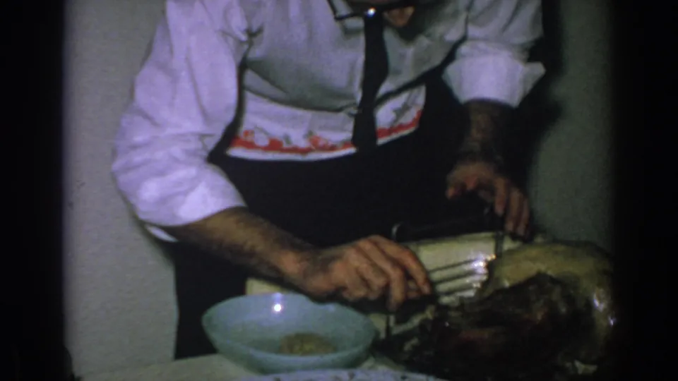 A man in a floral apron serves food with a spoon beside a bowl and dish in a dimly lit 1962 Gary, Indiana home kitchen.