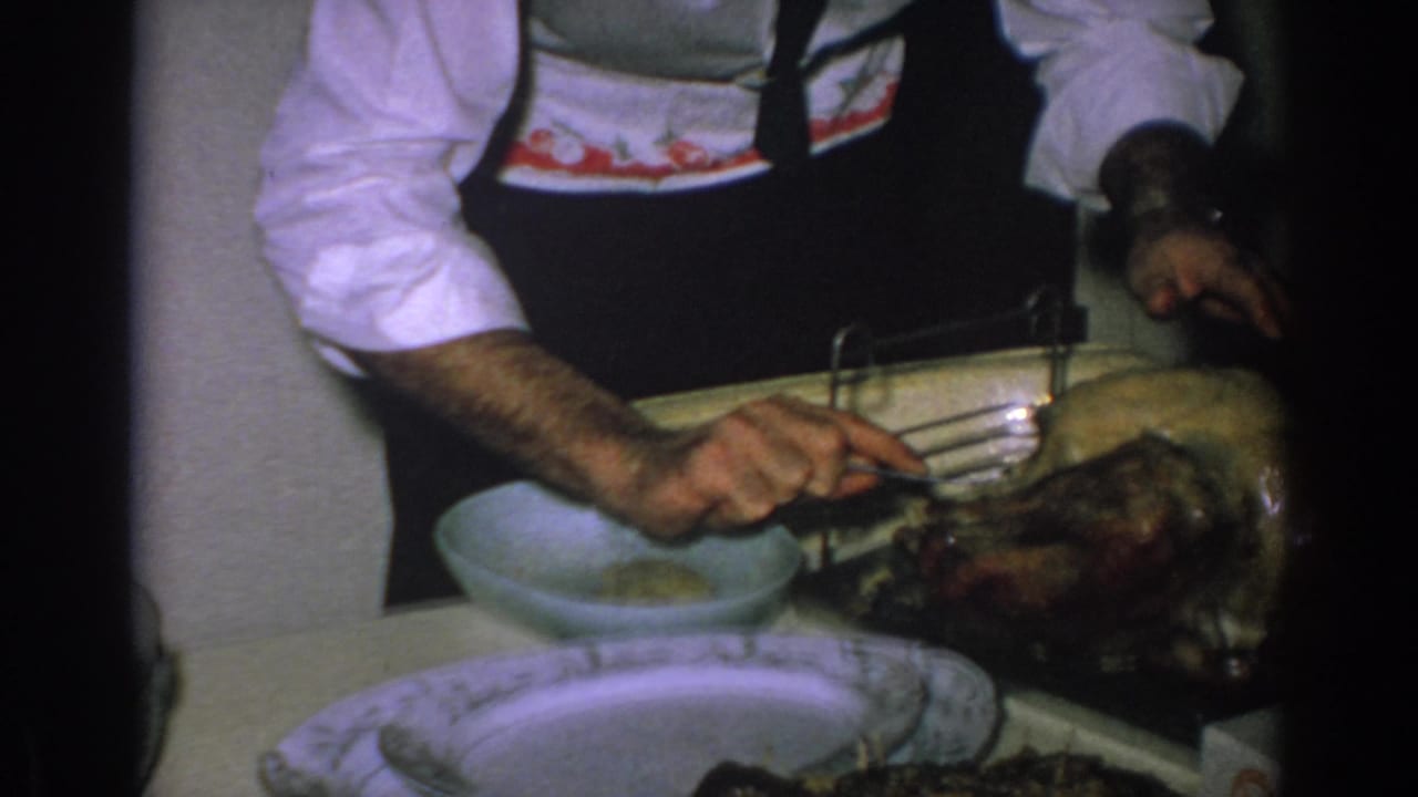 Man in white shirt and dark apron carving a roasted turkey at a kitchen table.