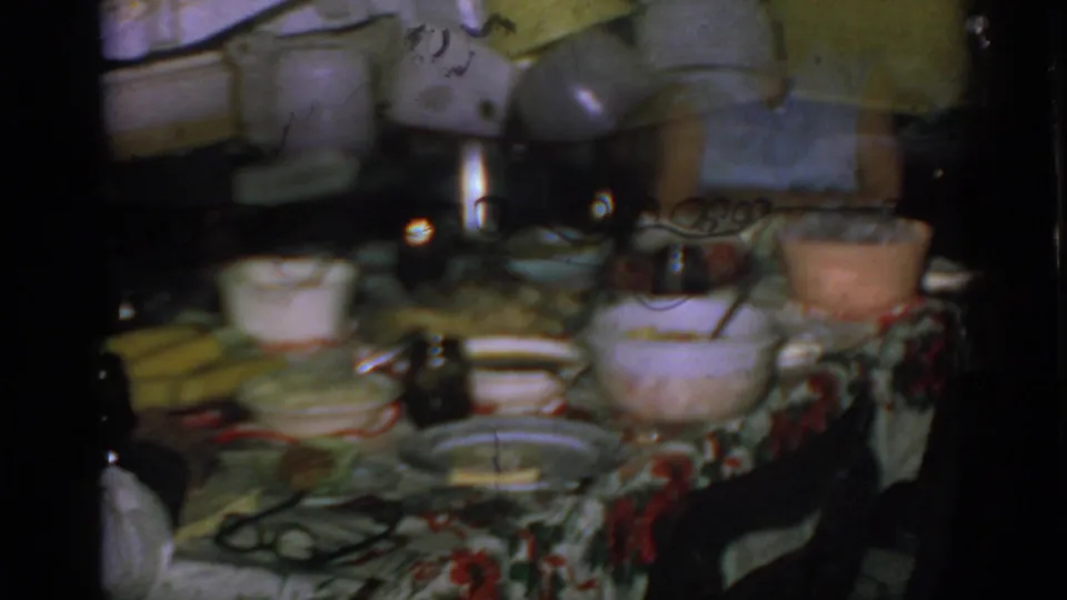 The camera turns to a crowded family dining table with serving bowls, plates, glasses, and a floral tablecloth inside the private home.