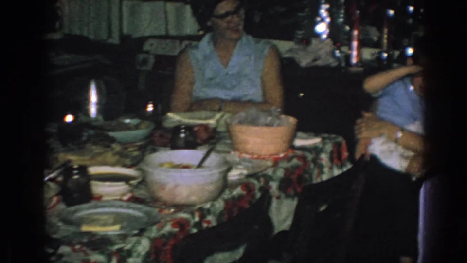 Adults sit around a meal table in the 1962 Gary, Indiana home-movie scene, with serving bowls, dishes, and dim interior lighting visible.