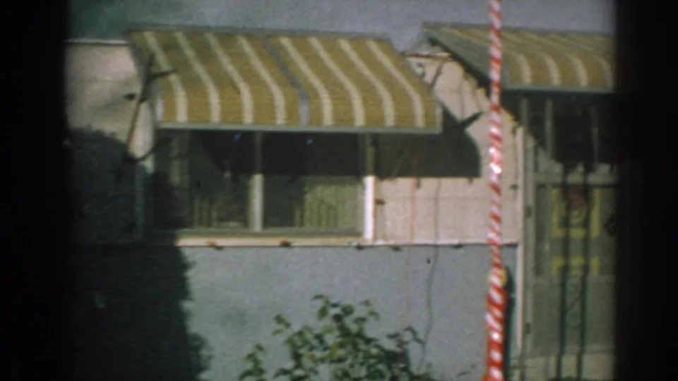 The camera view shifts along the blue trailer facade with striped awnings, windows, shrubs, and a striped holiday pole at the edge.