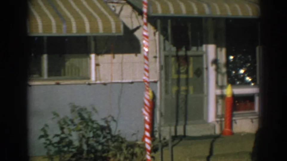 1962 home-movie frame of the trailer entrance with a glass-panel door, yellow-striped awning, red-and-white pole, shrubs, and red candle decoration.