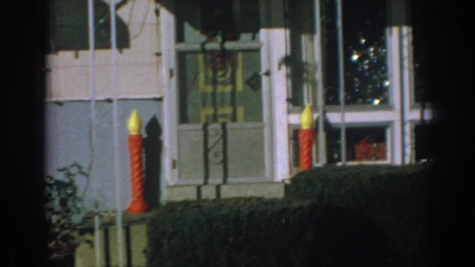 1962 Gary, Indiana home-movie frame of a front entrance with a red candle decoration, window, hedge, lawn, and partial candy cane ornament.