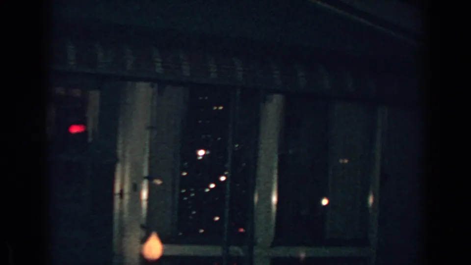 1962 Gary, Indiana home-movie frame showing a dim house exterior with a striped awning, dark window glass, small lights, and a warm blurred decoration near the bottom edge.