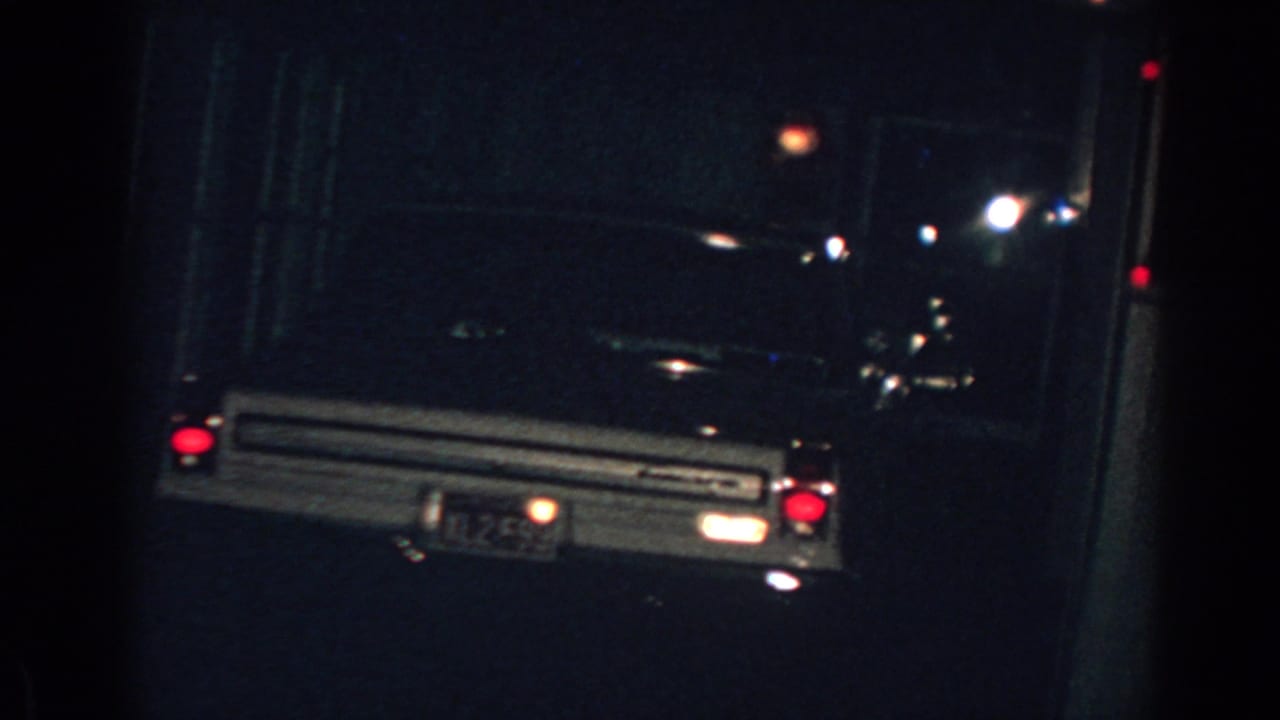 Parked car at night with red taillights visible beside a dark house exterior.