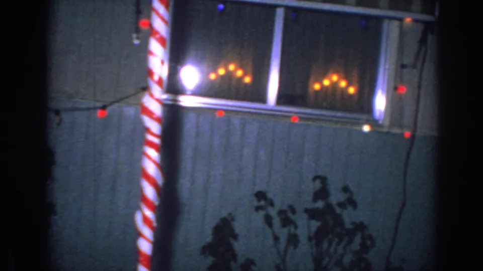 1962 Gary, Indiana frame of a house exterior at night with window lights, a red-and-white striped candy-cane prop, string lights, siding, and silhouetted plants.